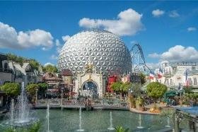 A large geodesic dome stands behind an amusement park area at Europa Park, featuring a fountain, buildings, greenery, and a rollercoaster under a blue sky with clouds.