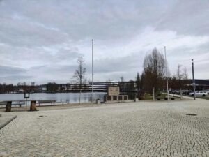 A paved square by a lake with bare trees, flagpoles, benches, and a monument; modern buildings and parked cars are visible in the background under a cloudy sky—a tranquil spot often visited during European Tours.